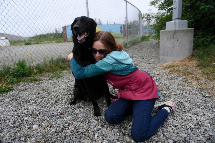 A woman kneels and hugs a large black dog by a fence
