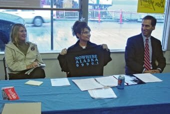Sarah Palin sits at a table between a man and a woman, holding up a t-shirt that says "Nowhere Alaska 99901"