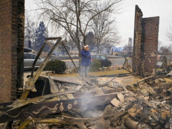 A woman stands with her hand over her mouth at the edge of the smoking ruins of a home.