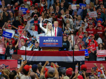 A woman in a white suit stands at a podium in a large crowd dressed in red, white and blue.