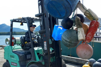 A man operates a forklift carrying a tangle of old buoys and a large plastic drum
