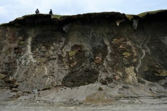 Two people standing atop a tall coastal bluff that is collapsing onto the beach