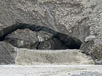 An ice cave mostly plugged by a huge chunk of ice, with water gushing out of it.