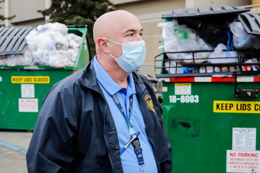 A bald man stands in front of two overflowing dumpsters