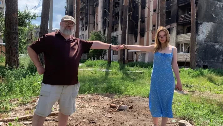 A middle-aged man and a young woman bump fists in front of a bombed out apartment building