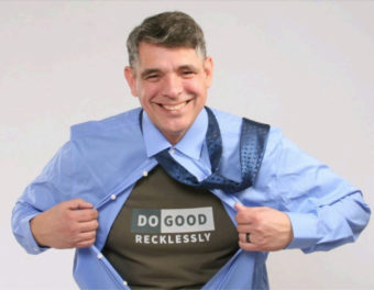 A grinning man tears open his button down shirt to reveal a t-shirt that says "do good recklessly" on it