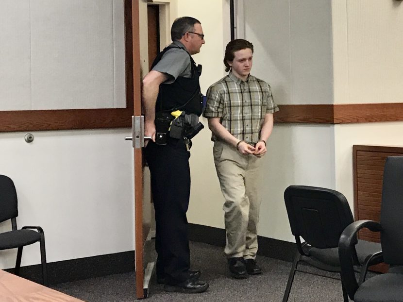 A bailiff holds a door open for a young man in handcuffs entering a courtroom