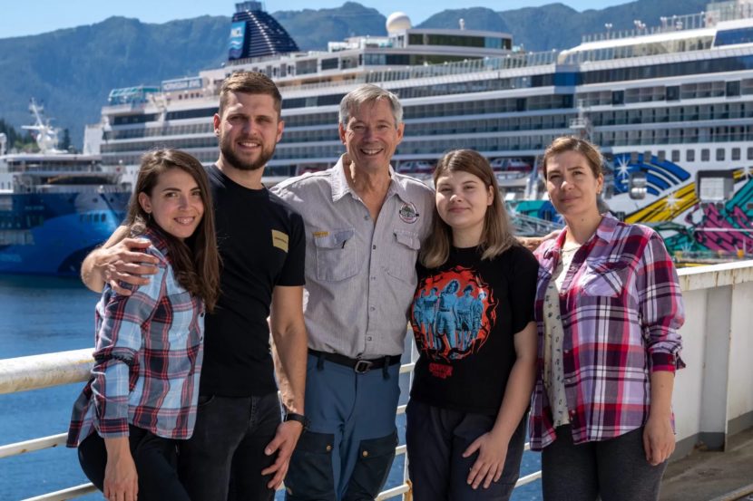 Five people pose on the deck of a ferry with a cruise ship behind them.