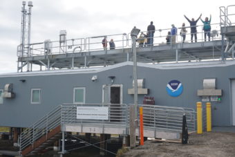 People stand and wave on top of a gray observatory building with the NOAA seal on it