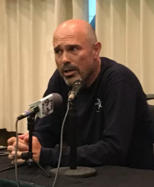A bald white man with a goatee speaks into a microphone