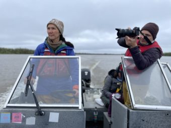 Peltola steers a boat while a cameraman stands next to her, filming