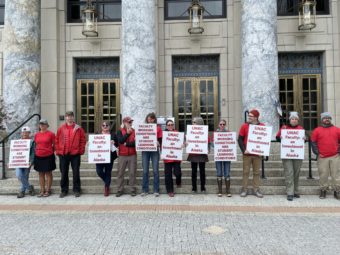 People stand in a row in front of the entrance to the Alaska Capitol holding signs supporting the faculty union