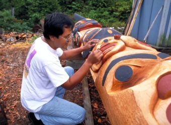 A man kneels and paints details on a totem pole lying on the ground