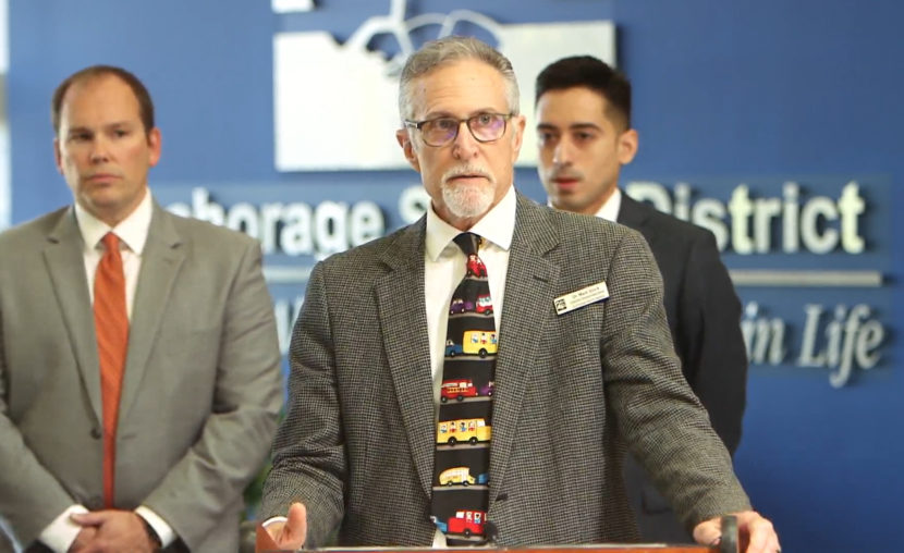Three men stand behind a lectern with stern expressions. One is wearing a tie with cartoon school buses on it.