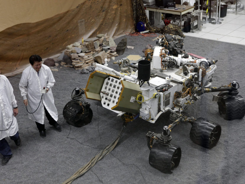 Scientists in lab coats stand around the Mars rover inside a building