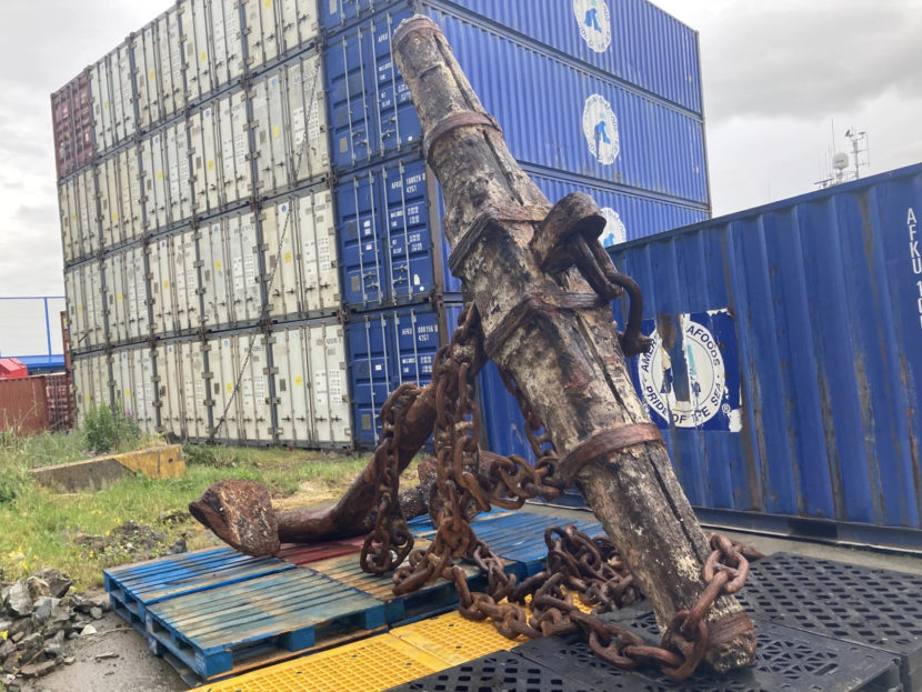 A big, rusty, old iron-and-wood anchor sitting on pallets next to a stack of shipping containers