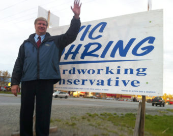 A man wearing a tie and a blue windbreaker stands waving in front of a large sign that says Vic Kohring, hardworking conservative