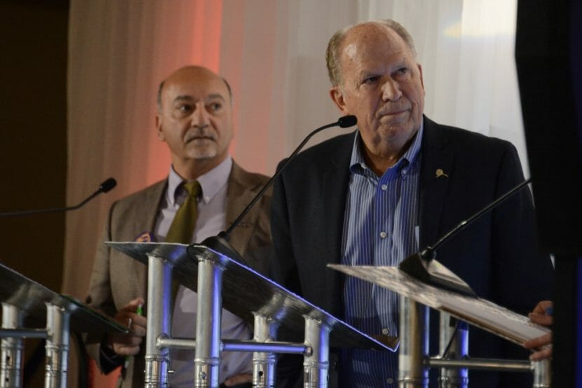 Bill Walker and Les Gara, both standing behind lecterns, appear to listen to someone off to their left