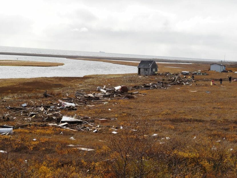 Plywood, lumber, and other debris strewn along a treeless coast, with a single small house in the background