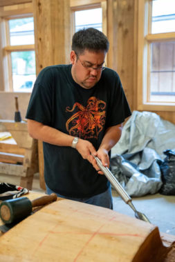 A man works on a log with a long wood chisel