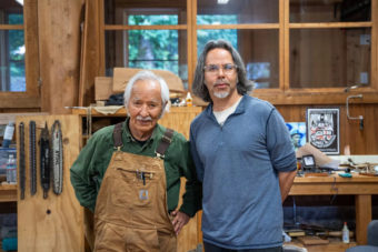 An older man and a younger man pose together in a woodshop