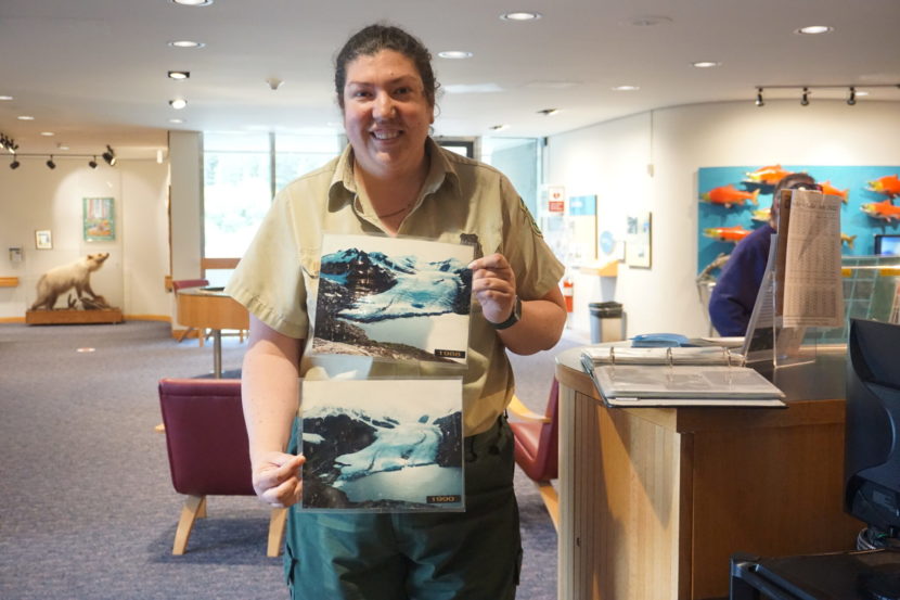 A woman stands in a visitors center holding two photographs demonstrating glacial retreat