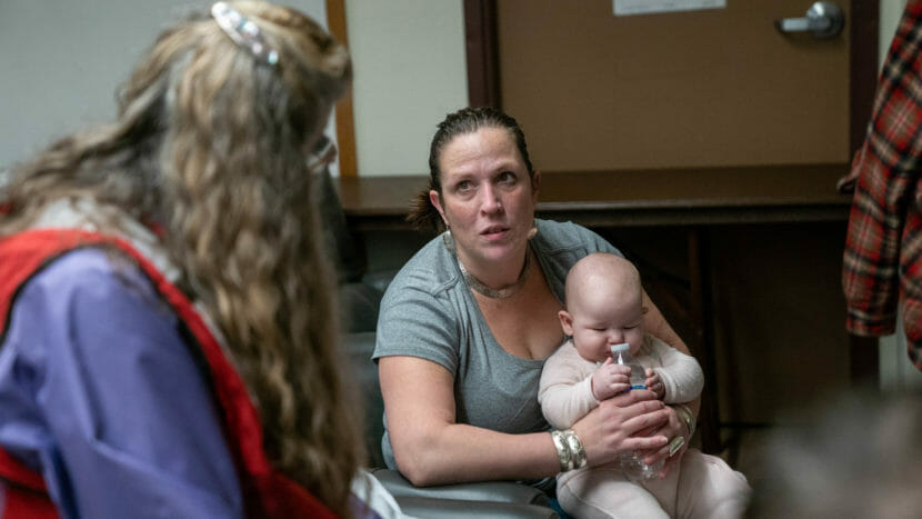 A woman holding a baby sits looking at a Red Cross volunteer, who is seen from behind