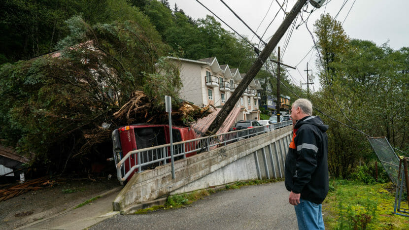 A man stands looking at a jumble of debris and a powerpole piled against a Toyota Tacoma lying on its side