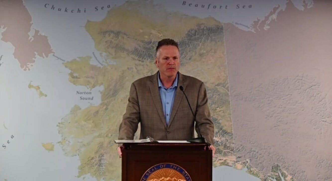 Gov. Dunleavy stands at a lectern in front of a map of Alaska
