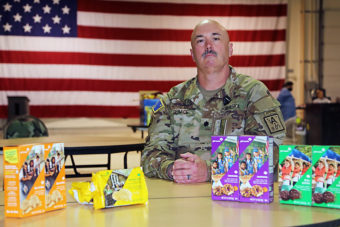 A bald man in uniform sits at a table covered with Girl Scout cookies, with an American flag behind him