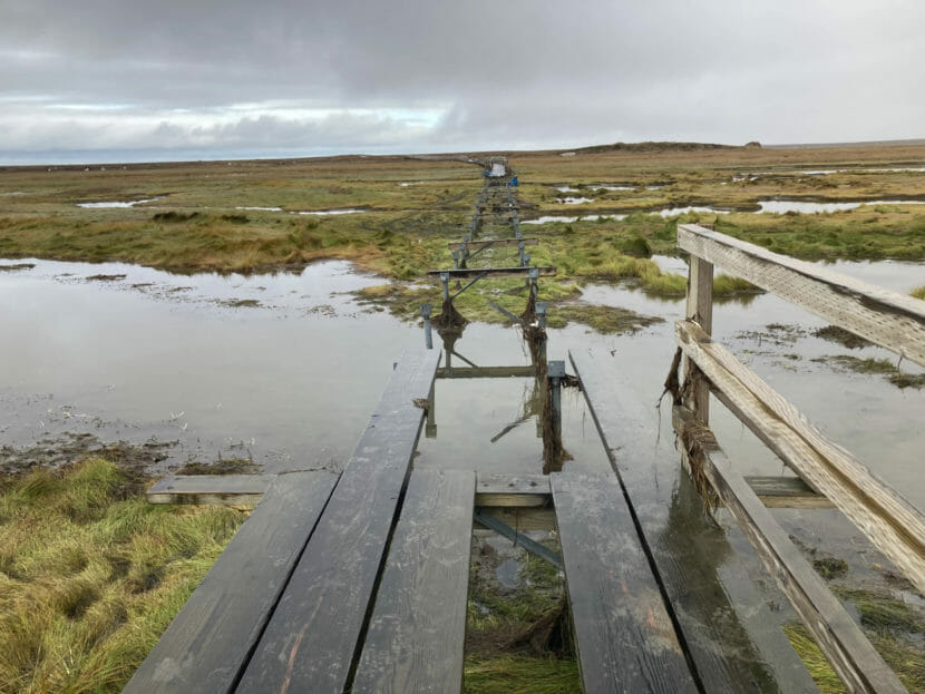 The frame of a boardwalk, with most of its planks gone, extending out into flooded tundra