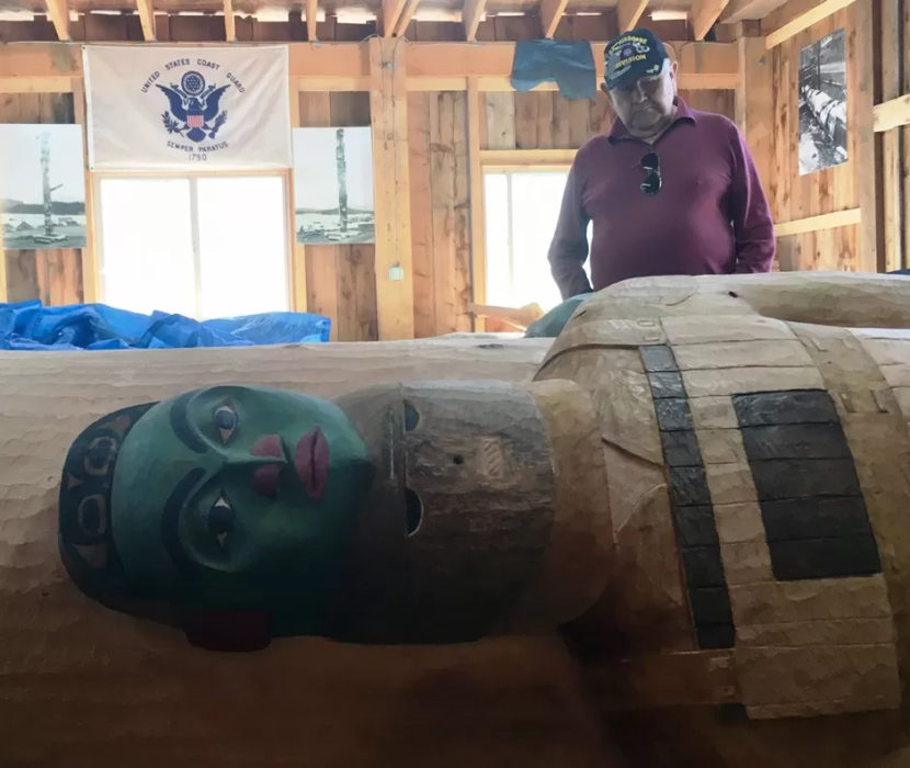 A man stands in a woodshop looking at totem pole lying on its side