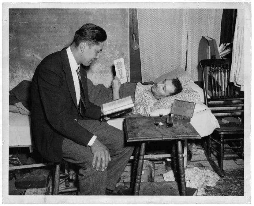 A man in a suit sits in a chair next to a man lying in bed