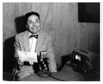 A black-and-white photo of a man in a suit and bowtie, sitting in front of a radio microphone