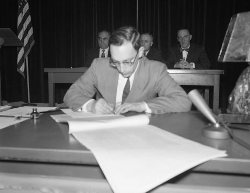 A black-and-white photo of a man in a suit signing a large document