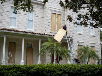 One man holes a ladder while another man lifts a quarter-sheet of plywood onto an awning roof
