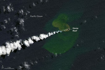 A satellite image shows a green volcanic island with a plume of white smoke coming out of a small black hole at its center.