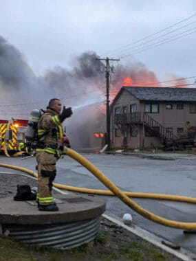 A man stands holding a firehose as a building burns across the street