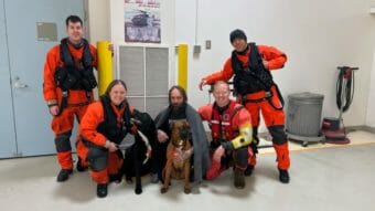 A soaking wet man, draped in a blanket, poses for a photo with four Coast Guard members and two dogs