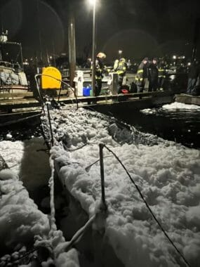 A nighttime photo showing firefighters on a dock, and firefighting foam covering what's left of a sailboat