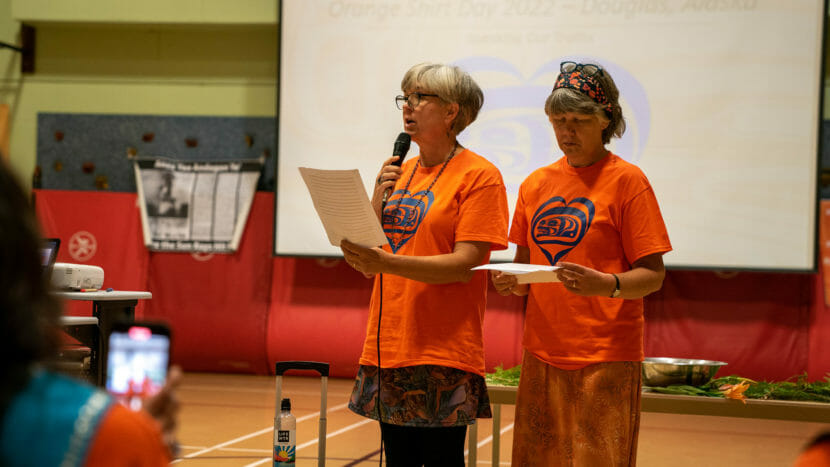 Two women wearing orange shirts stand and speak in a gymnasium