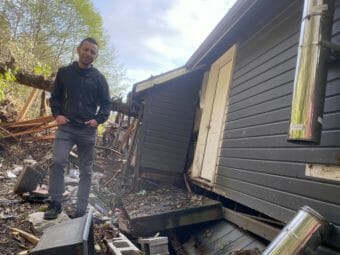 A man stands next to a tilty house that's off its foundation and impaled on a tree.