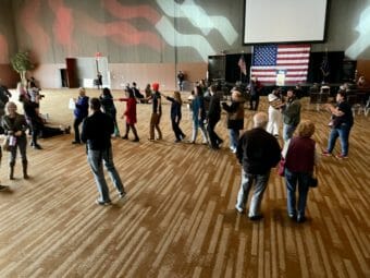 People standing around in a large, mostly empty hall. Some are walking in a line, but it's impossible to guess what they're doing.