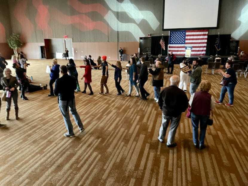 People standing around in a large, mostly empty hall. Some are walking in a line, but it's impossible to guess what they're doing.