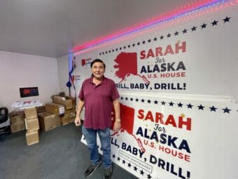 A man stands in a room with a giant Sarah Palin "drill baby drill" banner, next to a stack of cardboard boxes