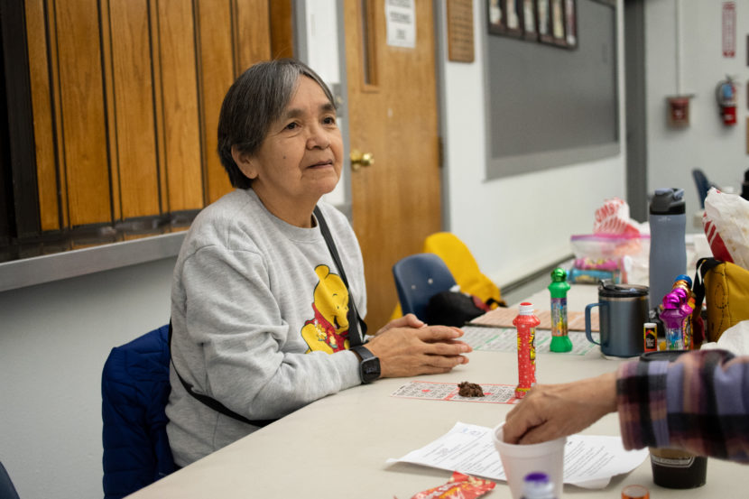 An older woman in a Winnie the Pooh sweatshirt sits at a folding table with bingo cards in front of her