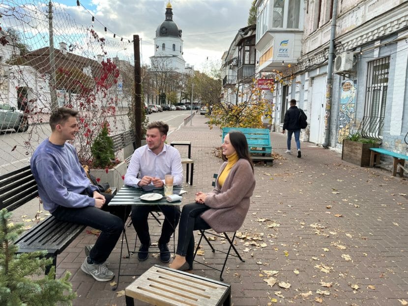 Three young people sit talking at an outdoor coffee shop table.