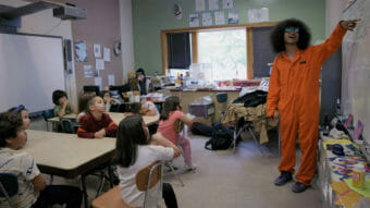 A man in an orange jumpsuit points at a whiteboard while schoolchildren look on