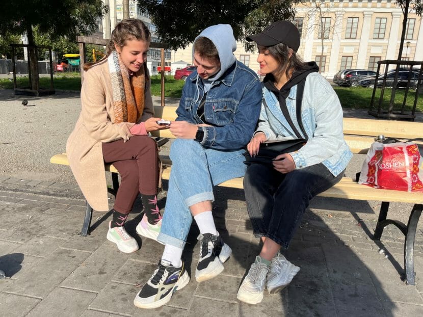 Three young people sit on a bench outside, looking at one of their phones