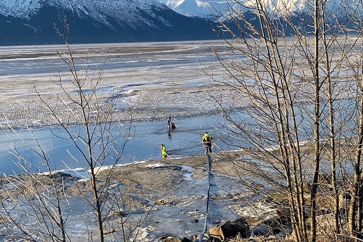 Seen from a distance, three rescuers work their way toward a man half-buried in coastal mud